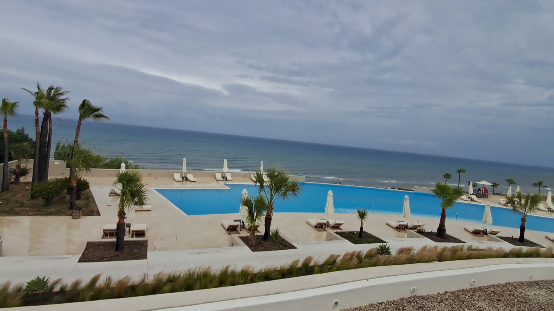 Grecotel La Riviera bezgalības baseins ar skatu uz Jonijas jūru mākoņainā dienā — sauļošanās krēsli un saulessargi piekrastē