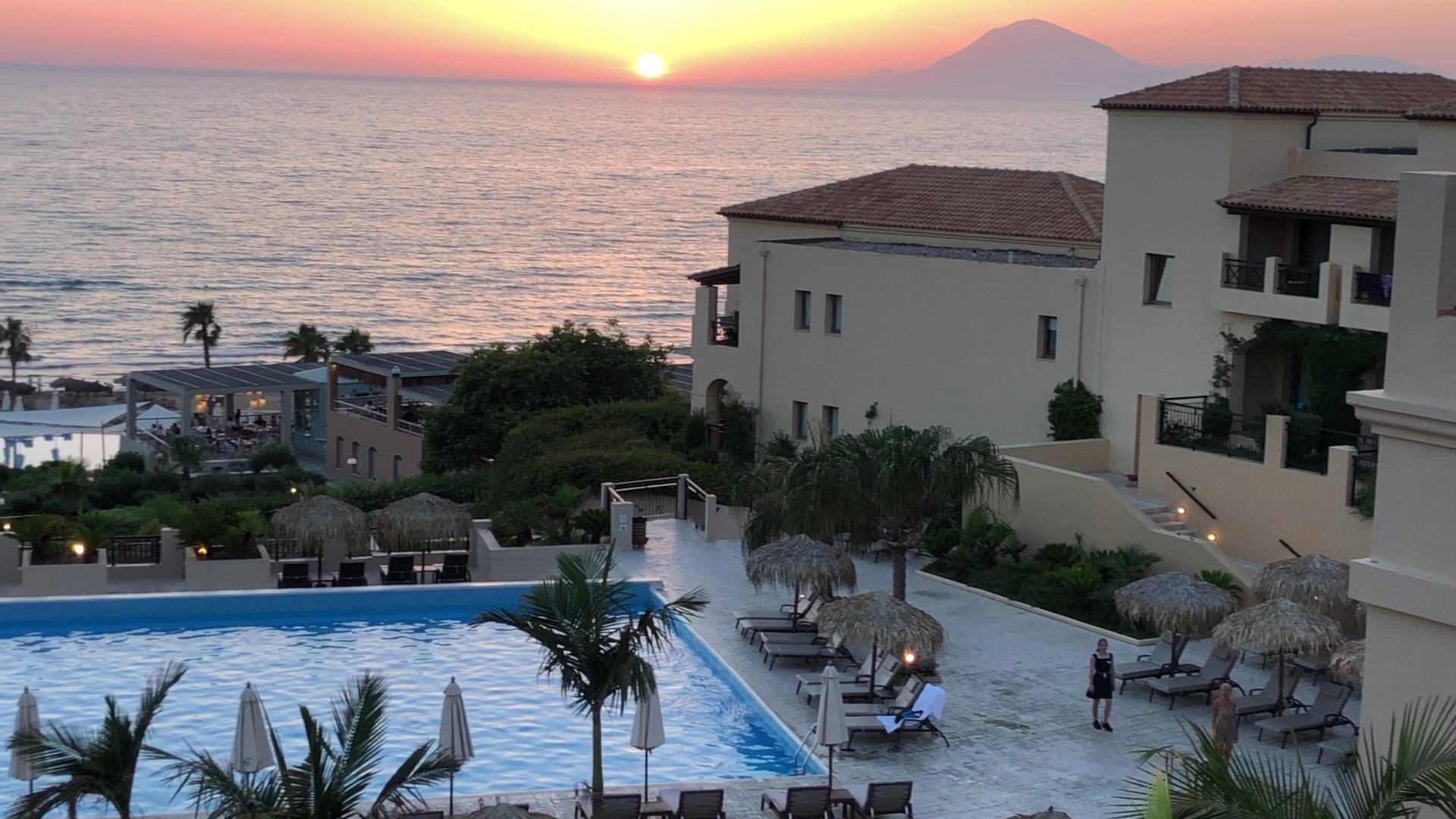 Saulriets virs Jonijas jūras pie Grecotel La Riviera viesnīcas ar skatu uz baseinu, palmām un kalnu tālumā