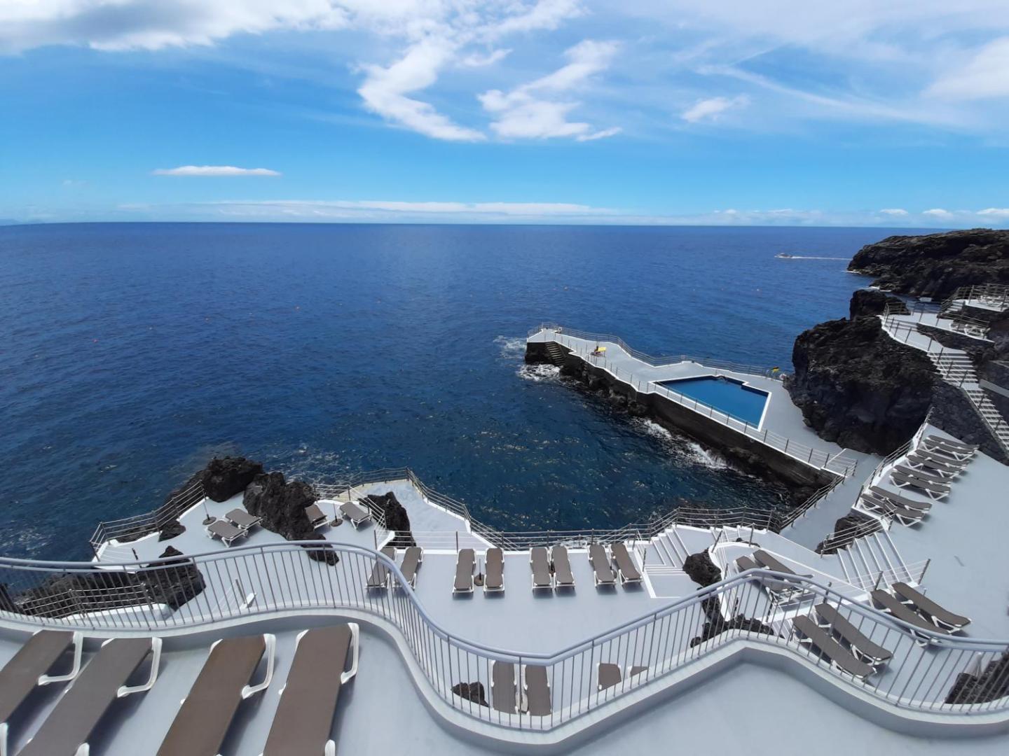 Panorāmas skats uz Rocamar Lido Resort atpūtas zonu pie okeāna ar sauļošanās krēsliem un nelielu baseinu uz klints