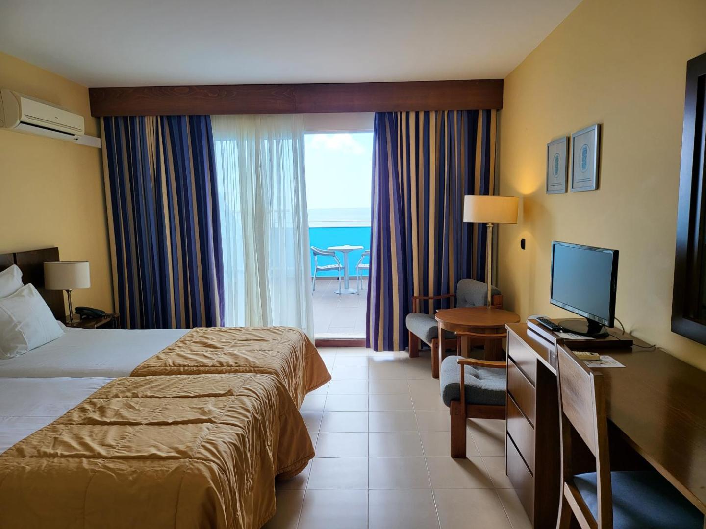 Viesnīcas Rocamar Lido Resort numura interjers ar rakstāmgaldu, televizoru un izeju uz privātu balkonu piekrastē