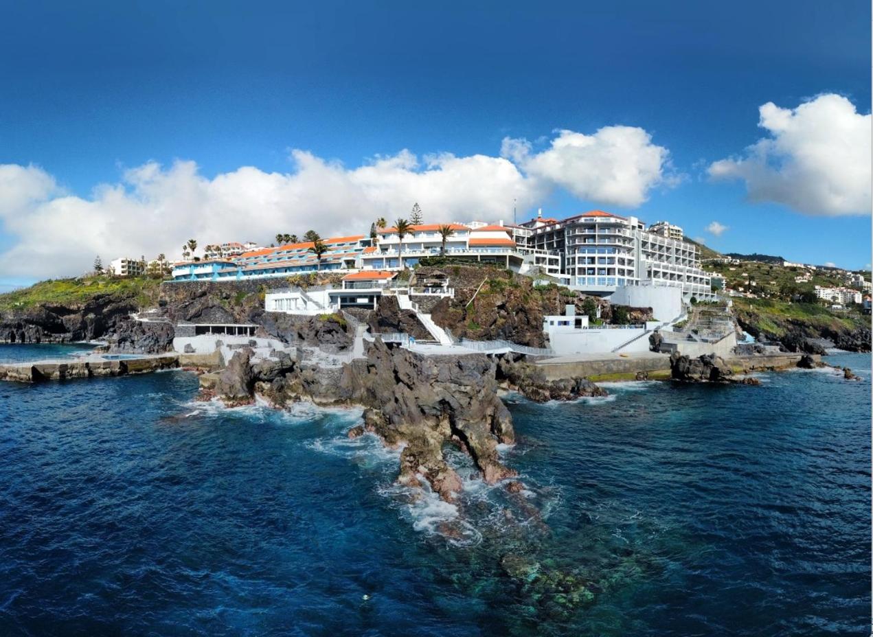 Rocamar Lido Resort panorāmas skats uz viesnīcas kompleksu Madeiras piekrastē — ēkas uz klints virs Atlantijas okeāna zilgmes