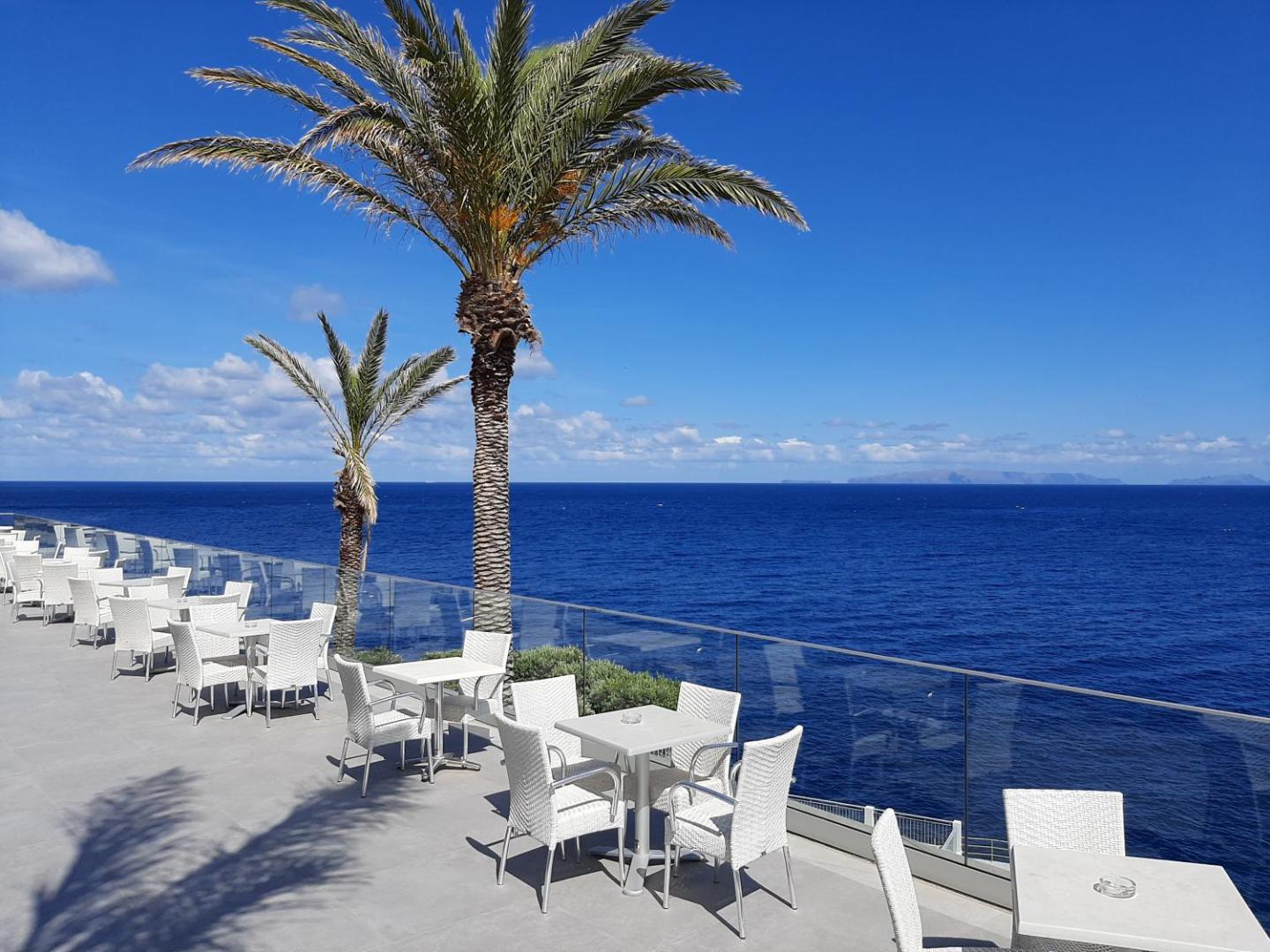 Rocamar Lido Resort saulainā āra terase ar baltām mēbelēm un palmām Madeiras piekrastē — panorāmas skats uz zilo okeānu