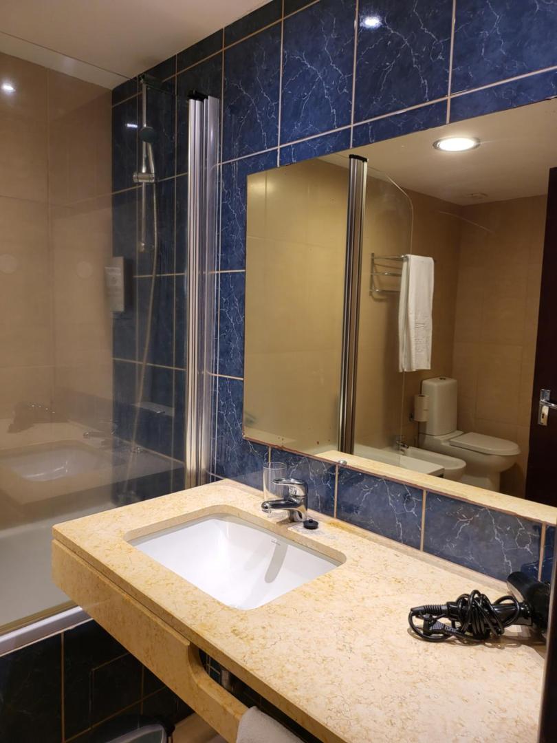 Rocamar Lido Resort numura vannas istaba ar zilu flīžu sienu, gaišu akmens virsmu, lielu spoguli un dušas zonu Madeirā