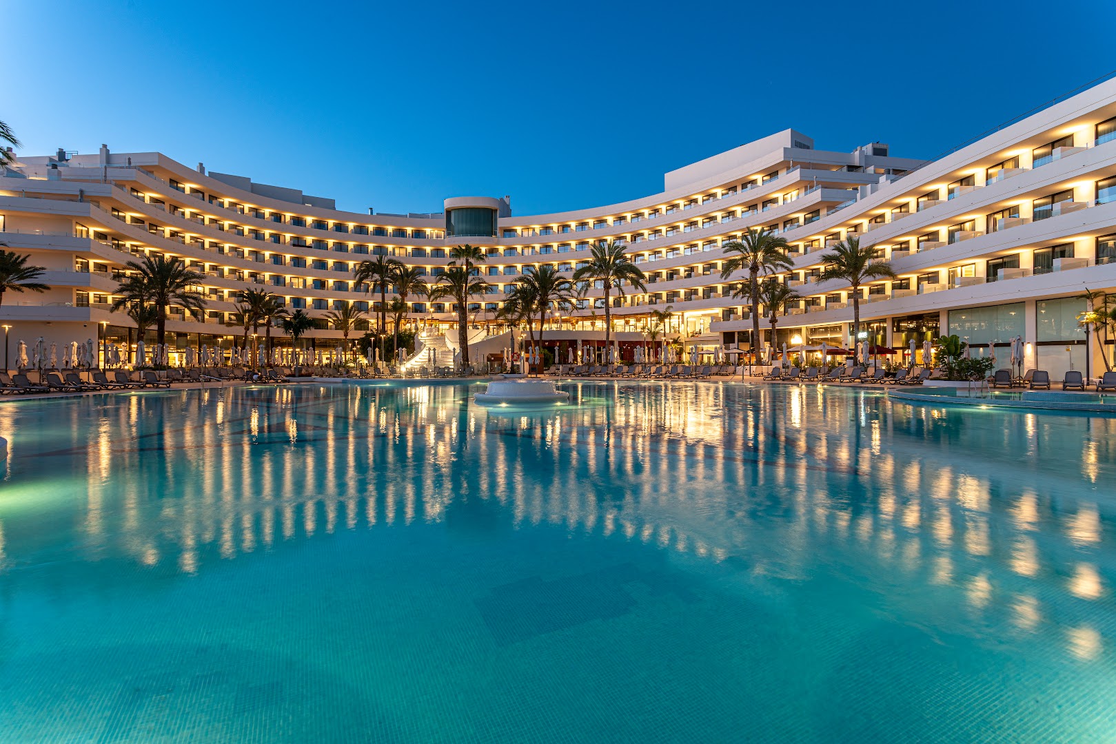 Skats uz Mediterranean Palace viesnīcas centrālo baseinu un balto arhitektūru zilos toņos vakara apgaismojumā Tenerifē.