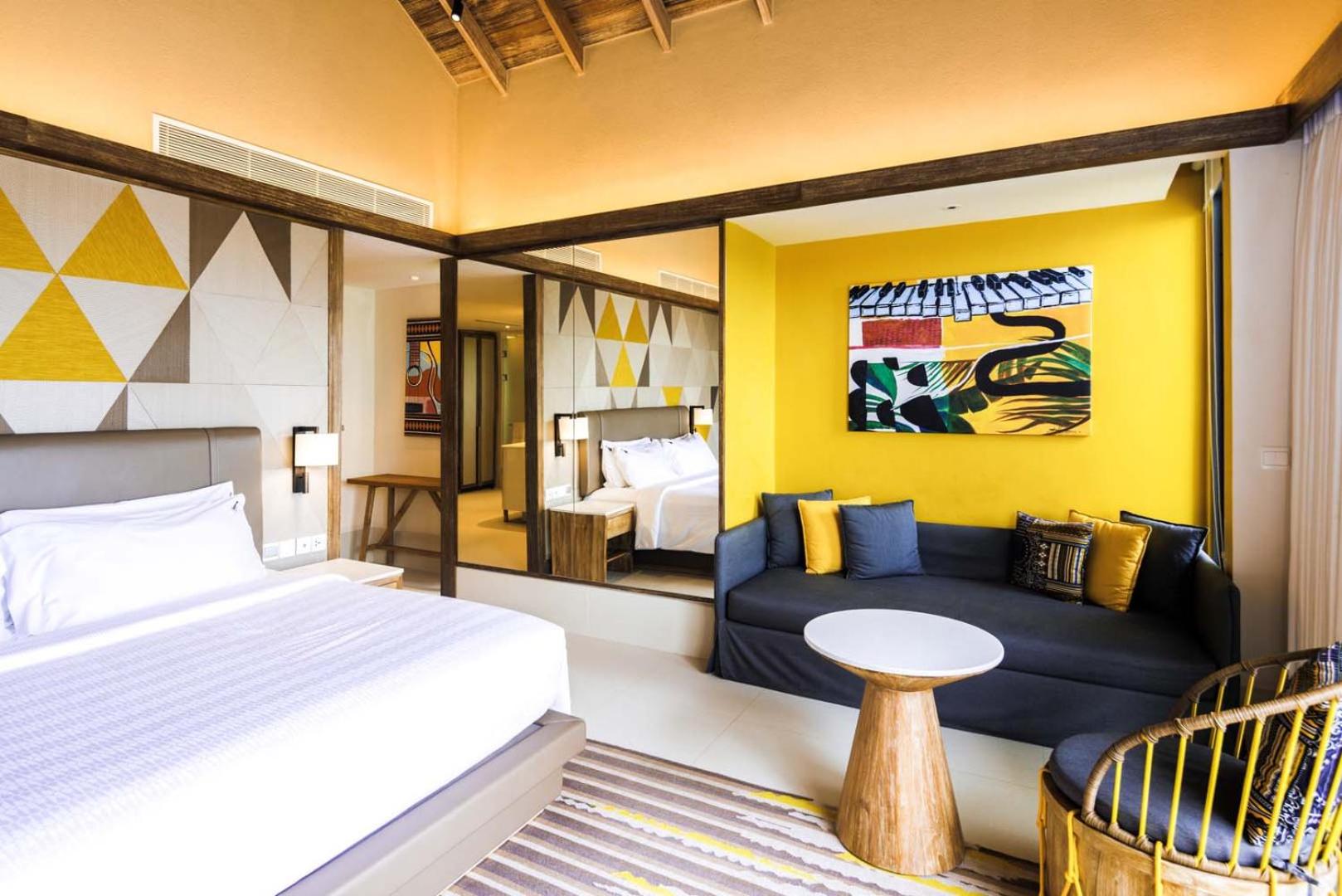 Hard Rock Hotel Maldives numura interjers ar lielu gultu, dzelteniem akcentiem pie sienām, dīvānu un stilīgu atpūtas krēslu.