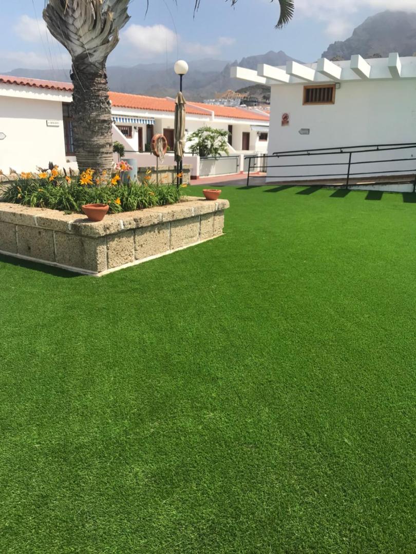 Malibu Park Apartments zaļā atpūtas zona ar mākslīgo zālienu, palmu un ziedošiem augiem uz kalnu fona Tenerifē
