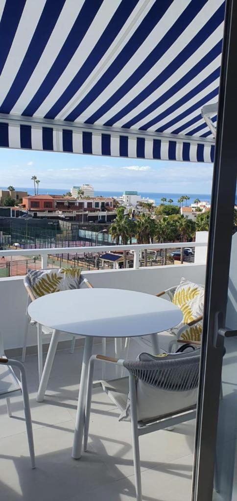 Skats no Malibu Park Apartments balkona uz okeānu un pilsētu — balts galds ar krēsliem zem svītrainas nojumes Tenerifē
