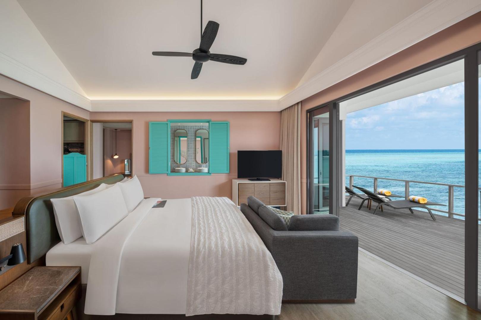 Le Meridien Maldives Resort & Spa guļamistaba ar privātu terasi un sauļošanās krēsliem tieši pie tirkīzzilā Indijas okeāna ūdens