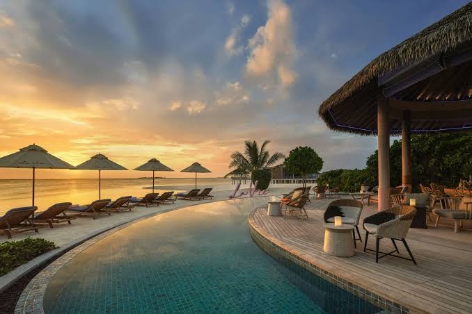 Le Meridien Maldives Resort & Spa saulrieta skats no āra terases ar atpūtas krēsliem pie bezgalības baseina okeāna krastā.