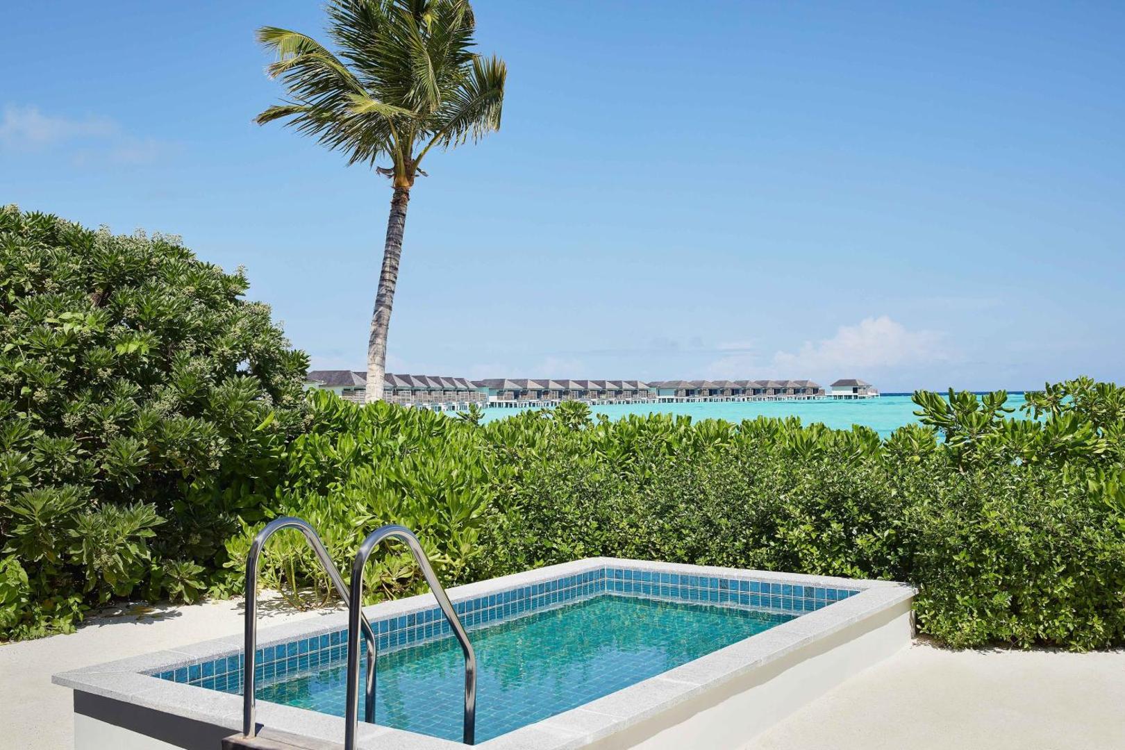 Le Meridien Maldives Resort & Spa neliels āra baseins dārzā ar skatu uz okeānu un ūdens bungalo tālumā zem skaidrām debesīm.