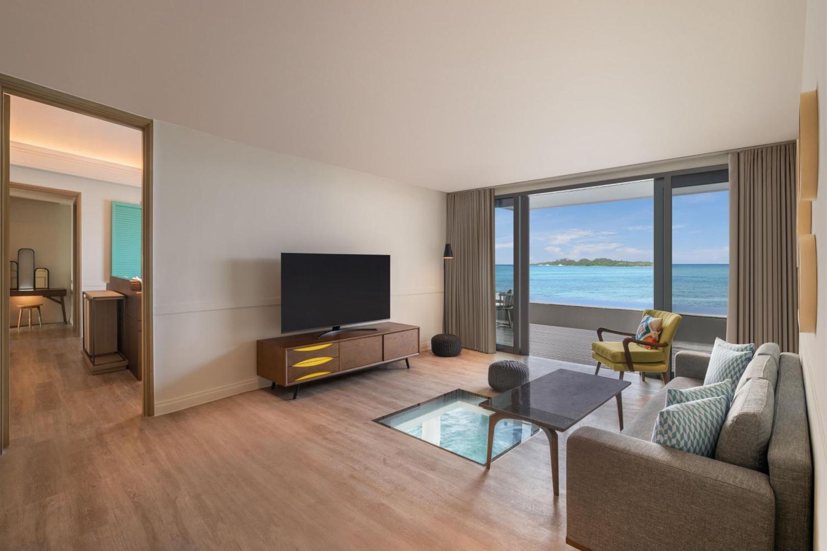 Le Meridien Maldives Resort & Spa viesistaba ar stikla grīdas logu, caur kuru redzams okeāns, un skatu uz piekrasti