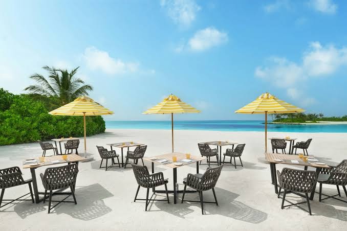 Le Meridien Maldives Resort & Spa pludmales restorāns ar dzelteniem saulessargiem un galdiņiem baltajās smiltīs pie okeāna