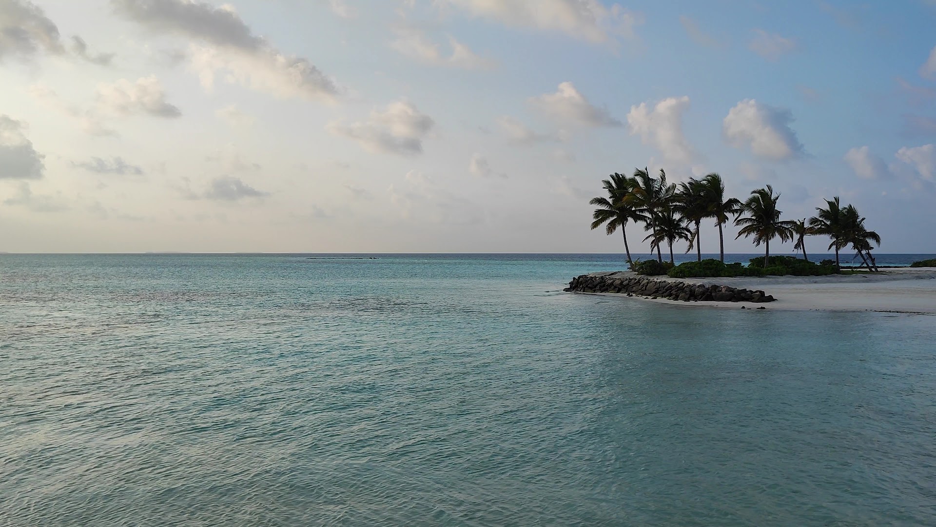 Le Meridien Maldives Resort & Spa skats uz tirkīzzilo okeānu un nelielu salu ar palmām un akmeņainu krastu saulrietā