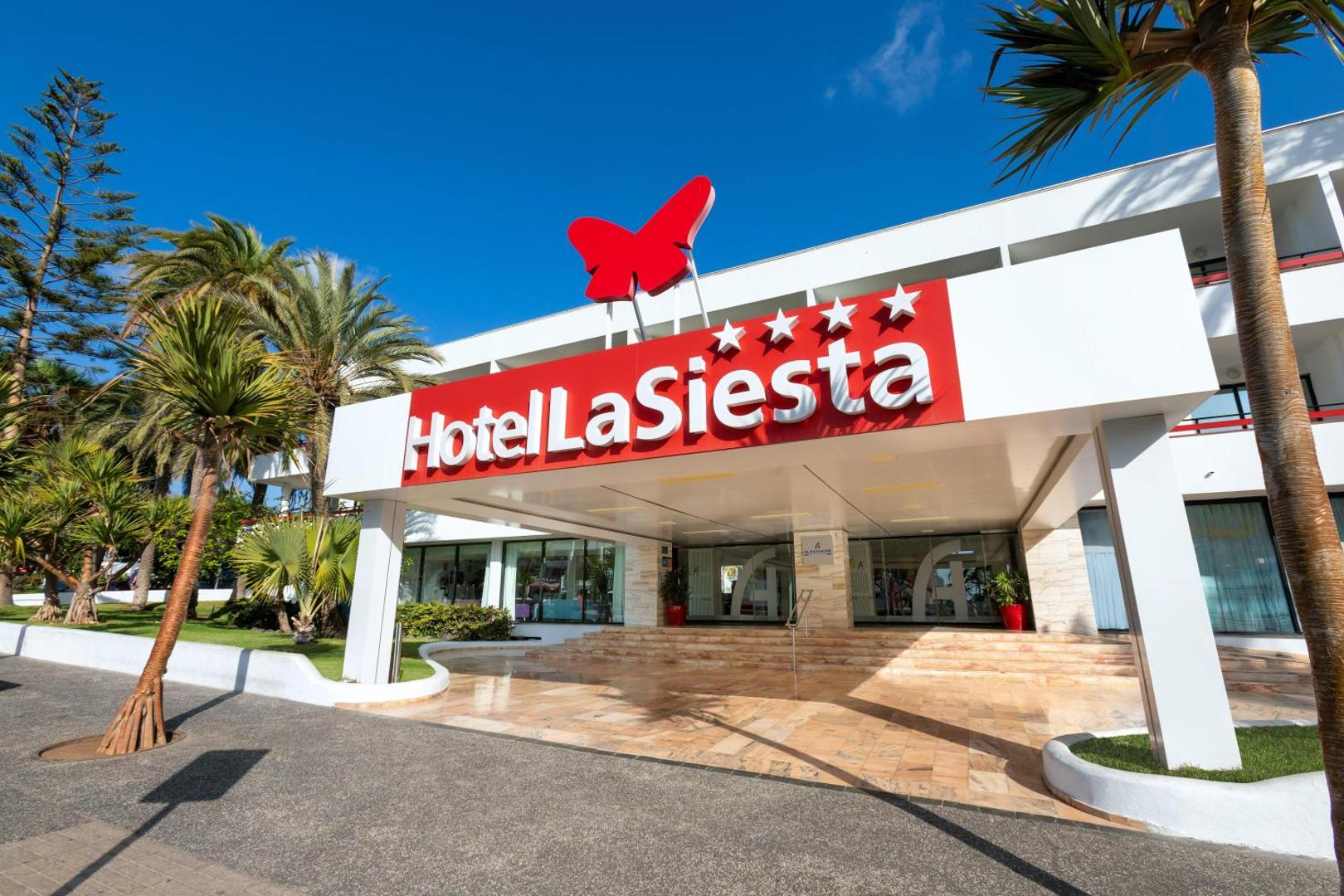 Alexandre Hotel La Siesta galvenā ieeja Playa de las Americas kūrortā Tenerifē — četru zvaigžņu viesnīcas fasāde ar palmām un zilu debesu fonu