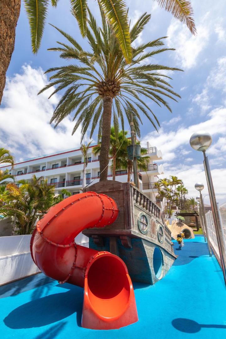 Alexandre Hotel La Siesta bērnu atrakcija kuģa formā ar sarkanu cauruļveida slidkalniņu zem palmām zilā debess fonā Tenerifē.