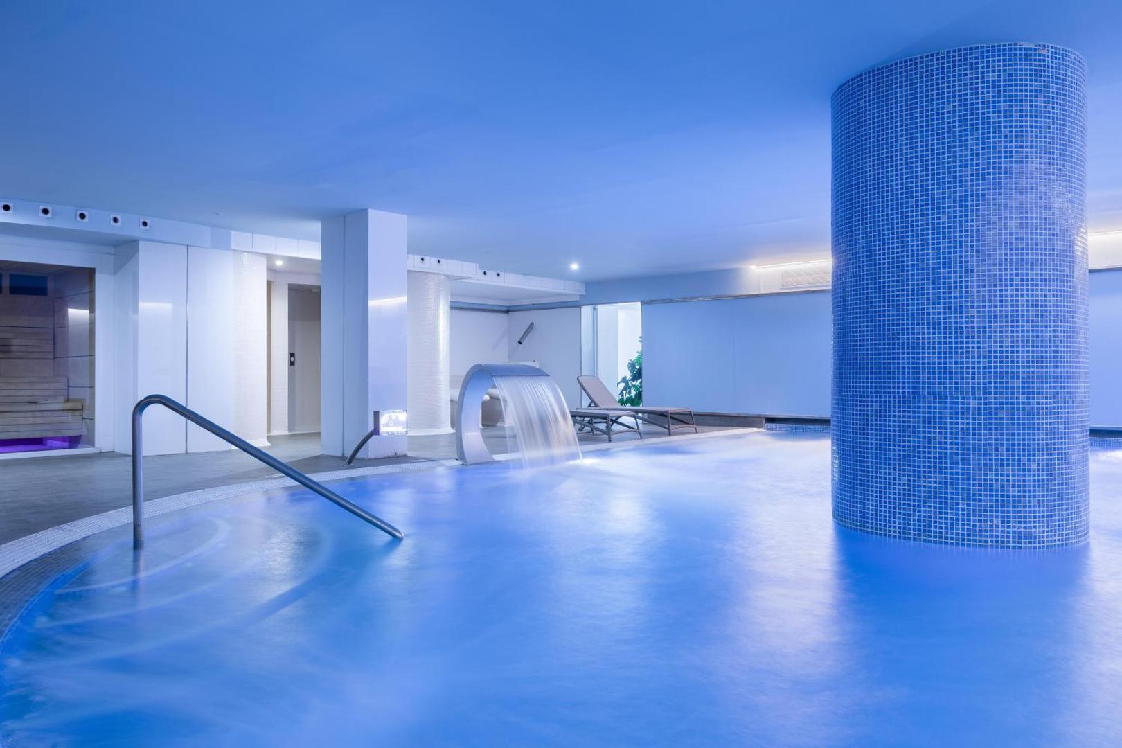 Alexandre Hotel La Siesta iekštelpu spa baseins ar zilu apgaismojumu, kaskādes ūdens strūklu un atpūtas krēsliem relaksācijas zonā