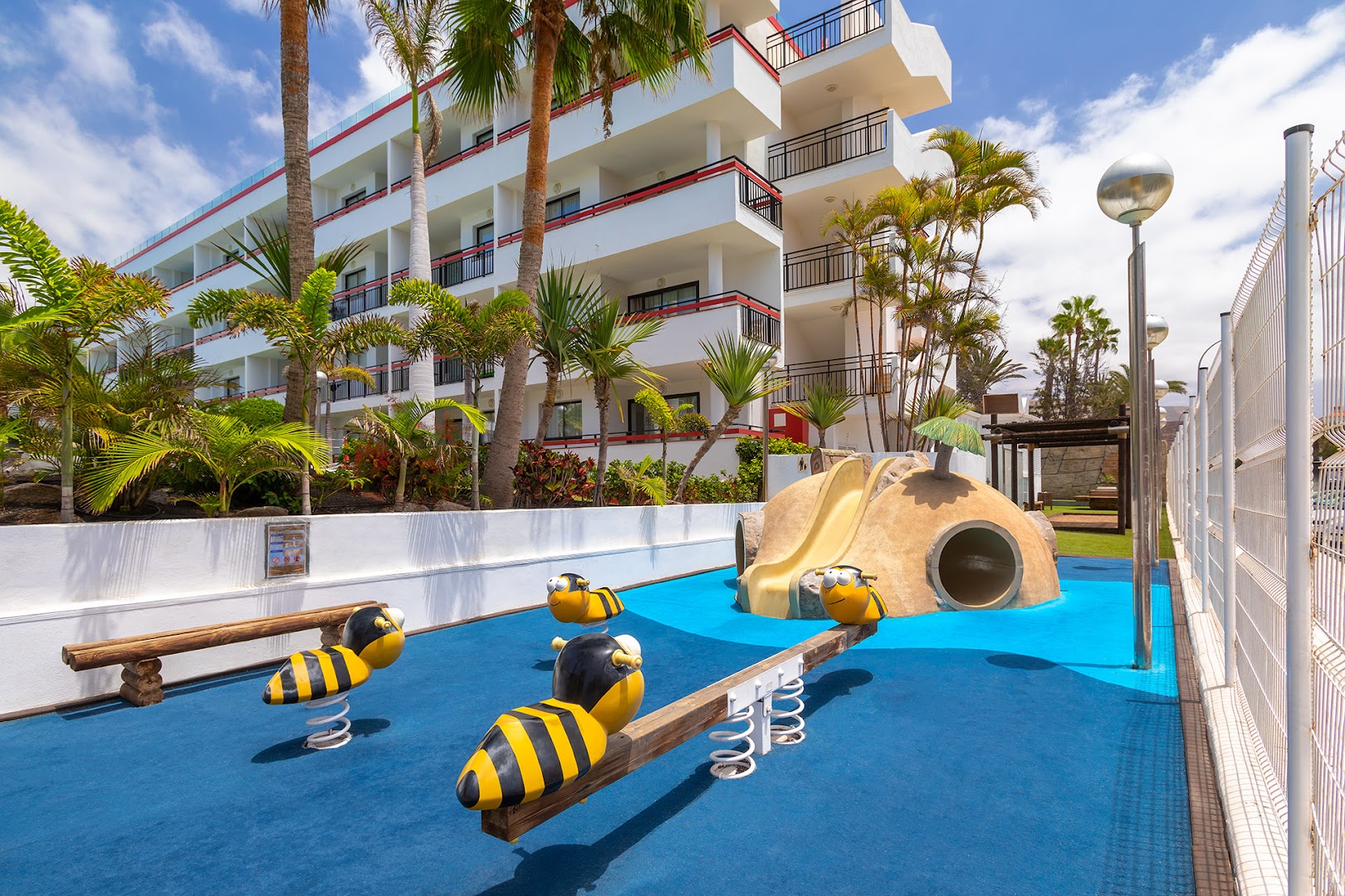 Alexandre Hotel La Siesta bērnu rotaļu laukums ar bitīšu šūpolēm un slidkalniņu pie viesnīcas korpusa Tenerifē, palmu ieskauts.