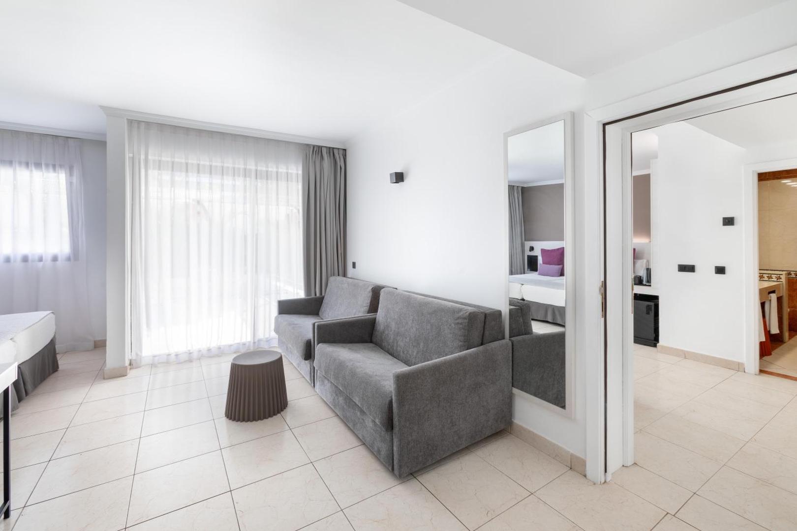 Alexandre Hotel La Siesta ģimenes numura viesistabas zona ar pelēku dīvānu, spoguli un ieeju guļamistabā un vannas istabā