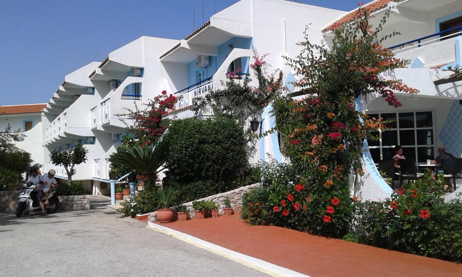 Nirvana Beach Hotel fasāde Rodas salā ar ziedošiem krūmiem, balkoniem un atpūtas zonu — tradicionāla Grieķijas arhitektūra baltos un zilos toņos.