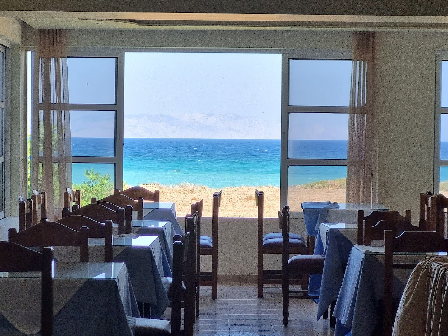 Nirvana Beach Hotel restorāna iekštelpas ar galdiņiem pie lieliem logiem, no kuriem paveras skats uz Egejas jūru
