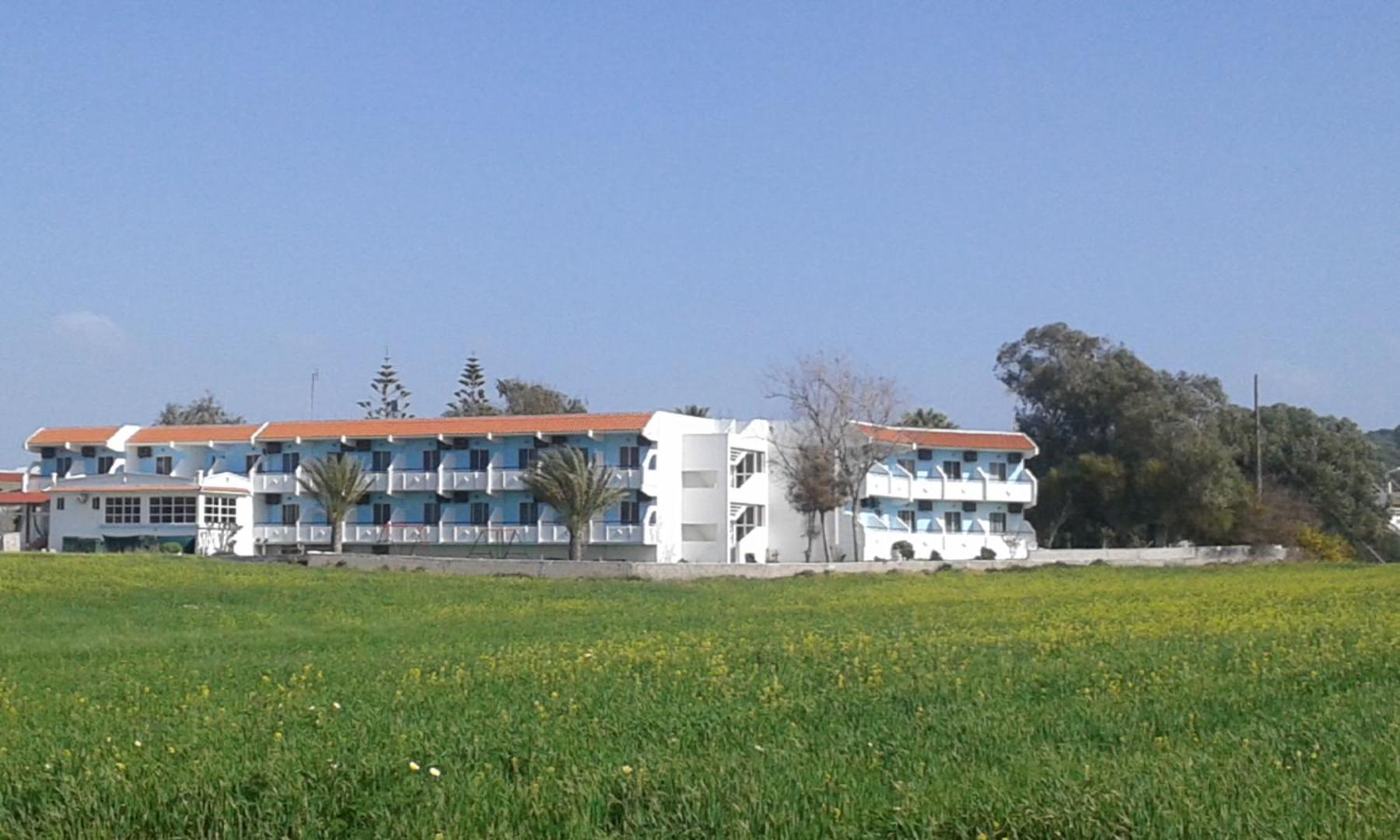 Nirvana Beach Hotel ēkas kopskats no blakus esošā lauka Rodas salā — trīs stāvu viesnīca ar sarkaniem dakstiņu jumtiem un palmām.