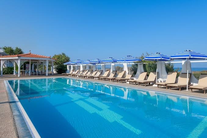 Nirvana Beach Hotel āra baseins Rodas salā ar sauļošanās krēsliem, zili svītrainiem saulessargiem un koka lapeni fonā zem skaidrām debesīm