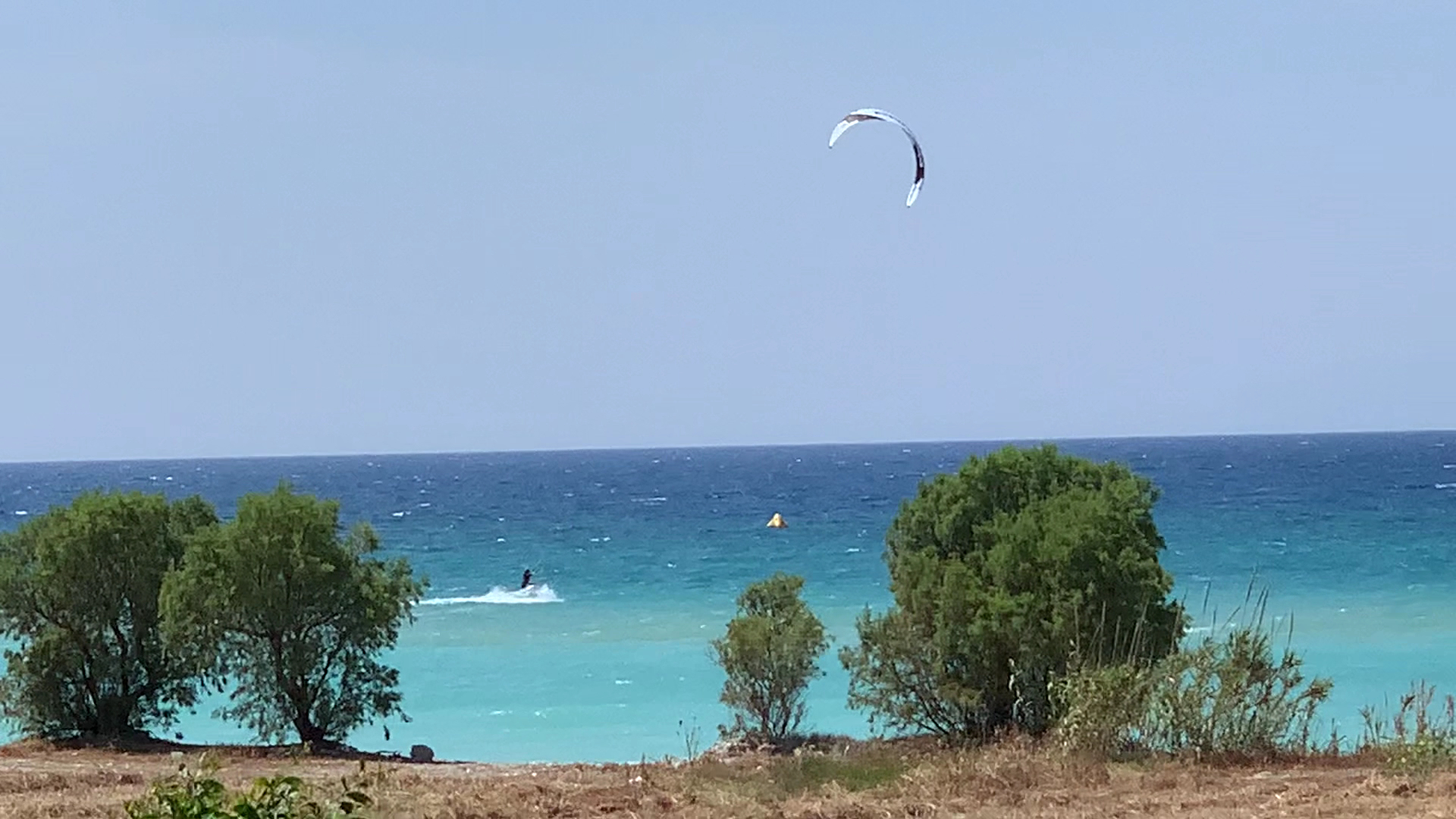 Nirvana Beach Hotel skats uz Egejas jūru Rodas salā, kur redzams kaitbordists un piekrastes koki zilā ūdens fonā