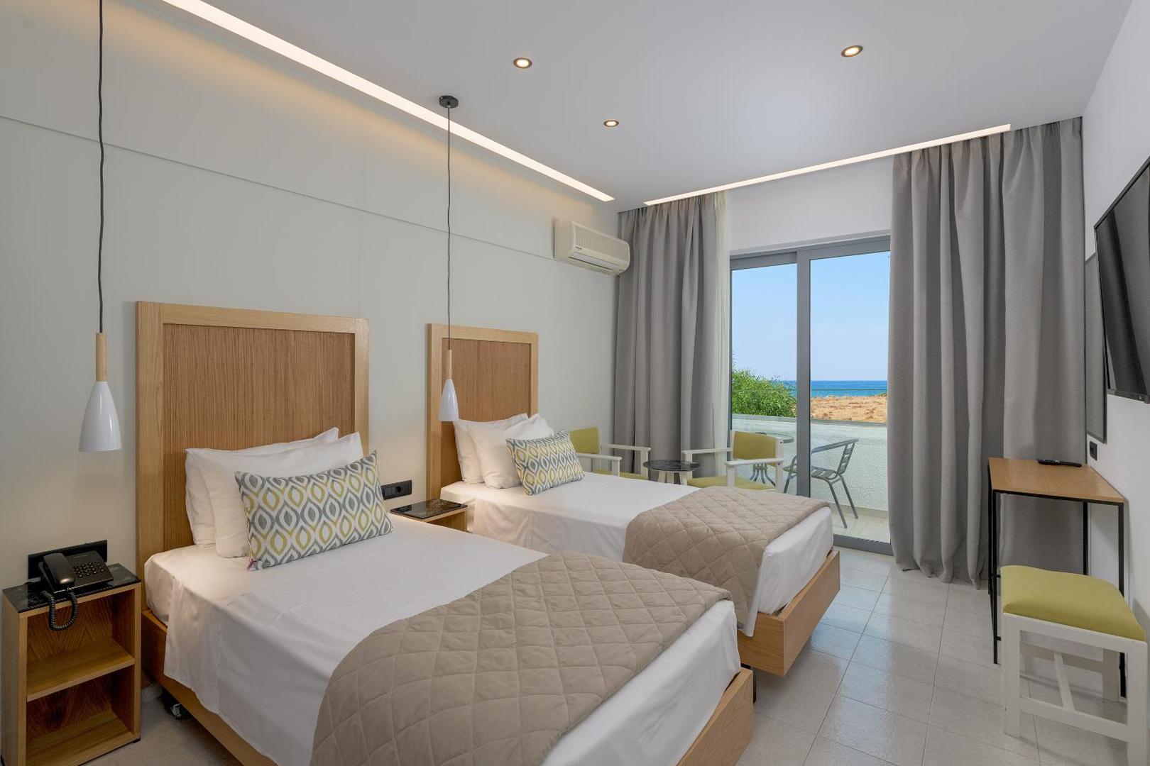 Moderns Nirvana Beach Hotel divvietīgs numurs ar gaišām mēbelēm, divām gultām un lielu logu, kas ved uz balkonu ar skatu uz jūru.