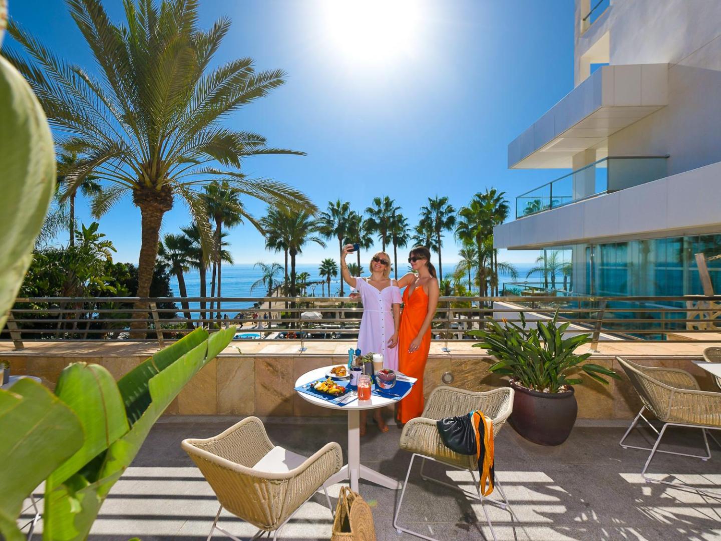 Sievietes uz Amare Beach Hotel Marbella terases bauda brokastis ar skatu uz Vidusjūru un palmām saulainā laikā.