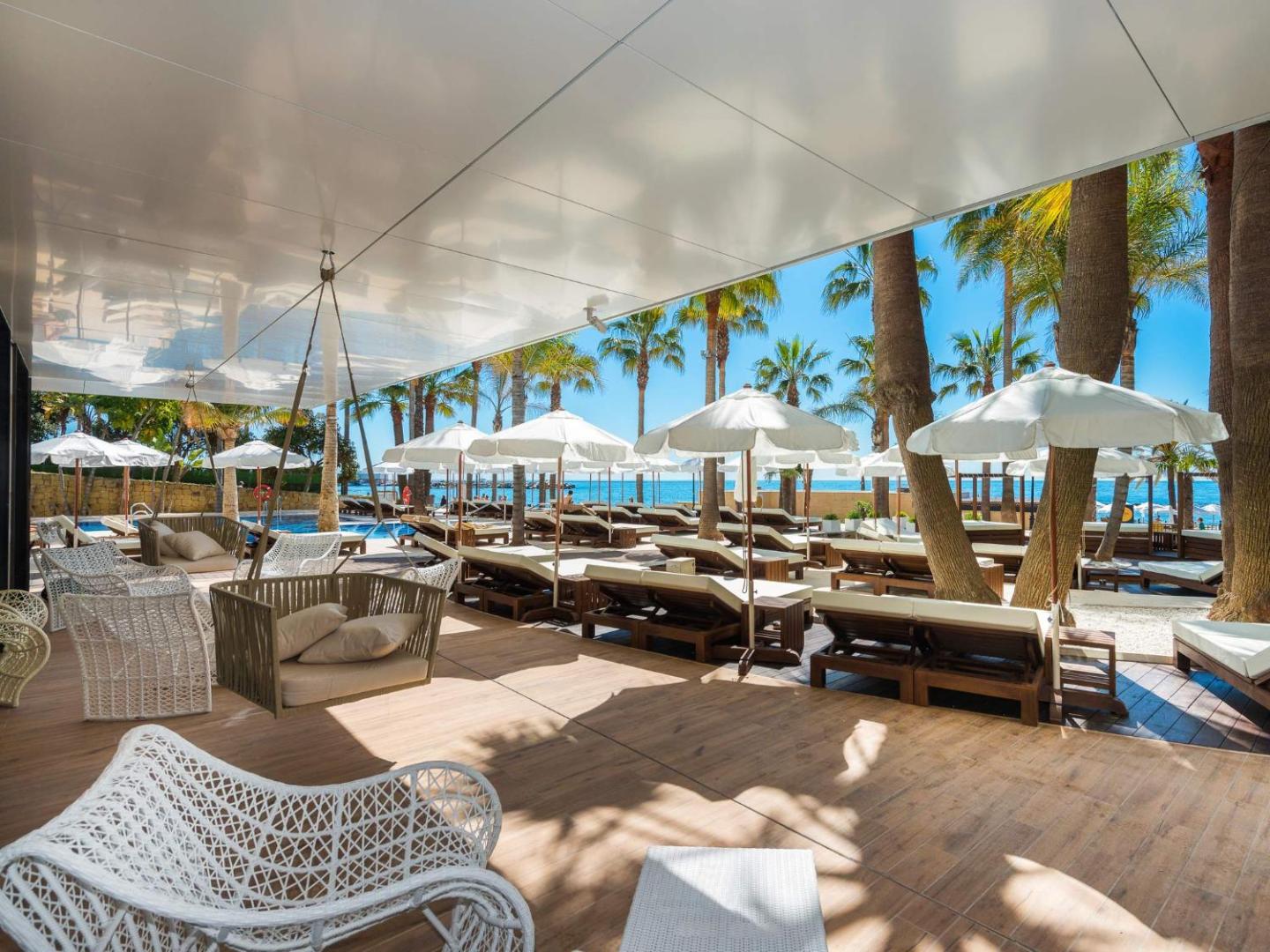 Amare Beach Hotel Marbella atpūtas zona pie pludmales ar šūpuļkrēsliem, sauļošanās krēsliem un saulessargiem palmu ielokā Vidusjūras krastā.