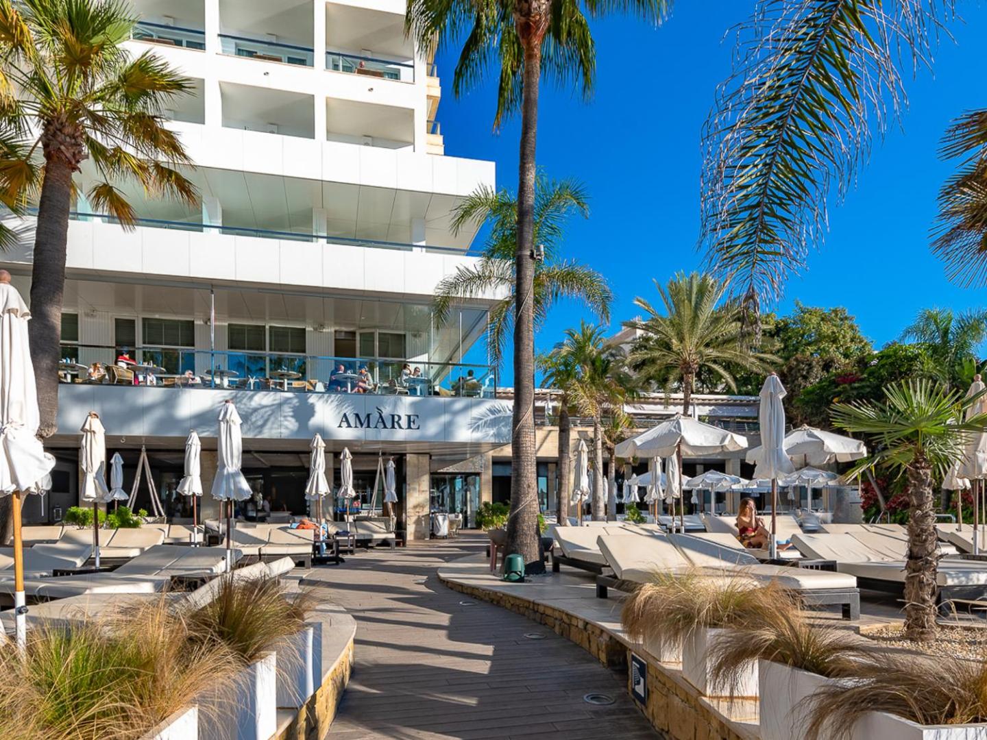 Amare Beach Hotel Marbella āra terase ar atpūtas krēsliem un saulessargiem palmu ēnā pie viesnīcas fasādes Marbeljā, Spānijā