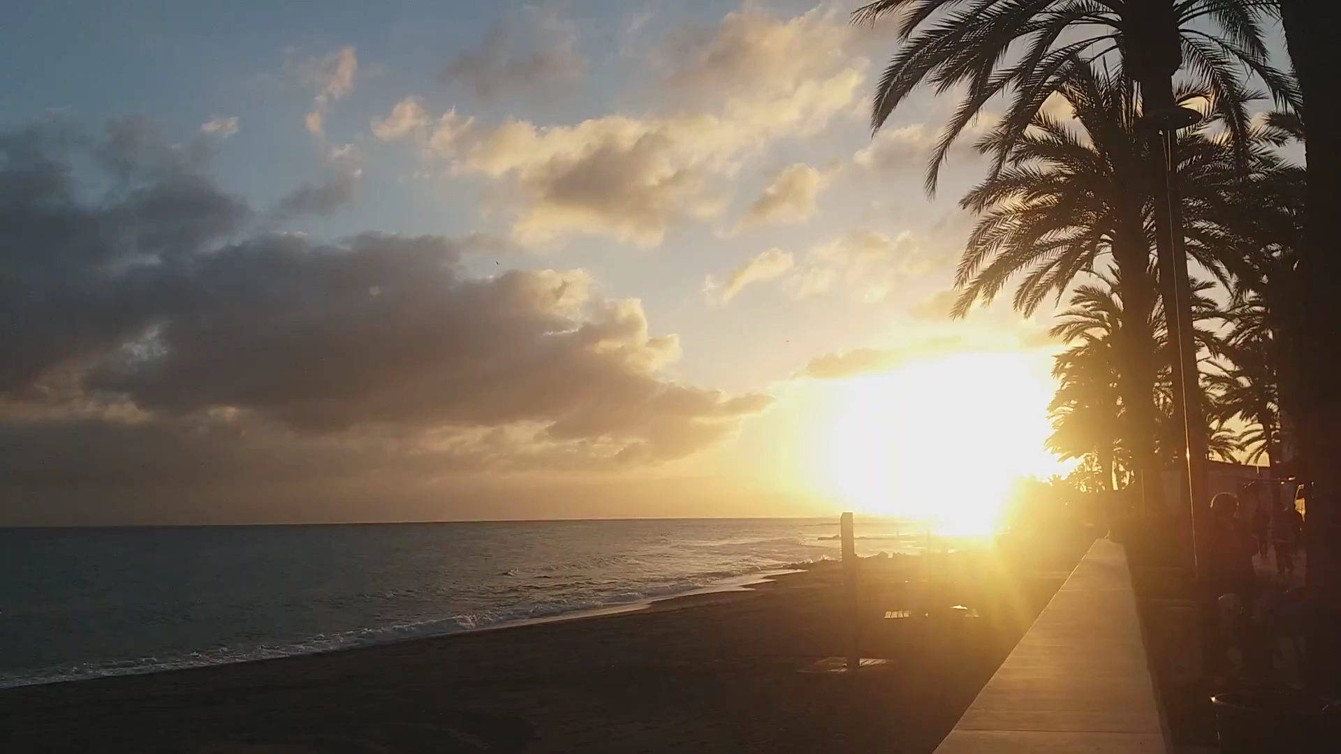 Saulriets Amare Beach Hotel Marbella pludmalē ar palmu siluetiem un saules atspulgu jūrā pie horizonta