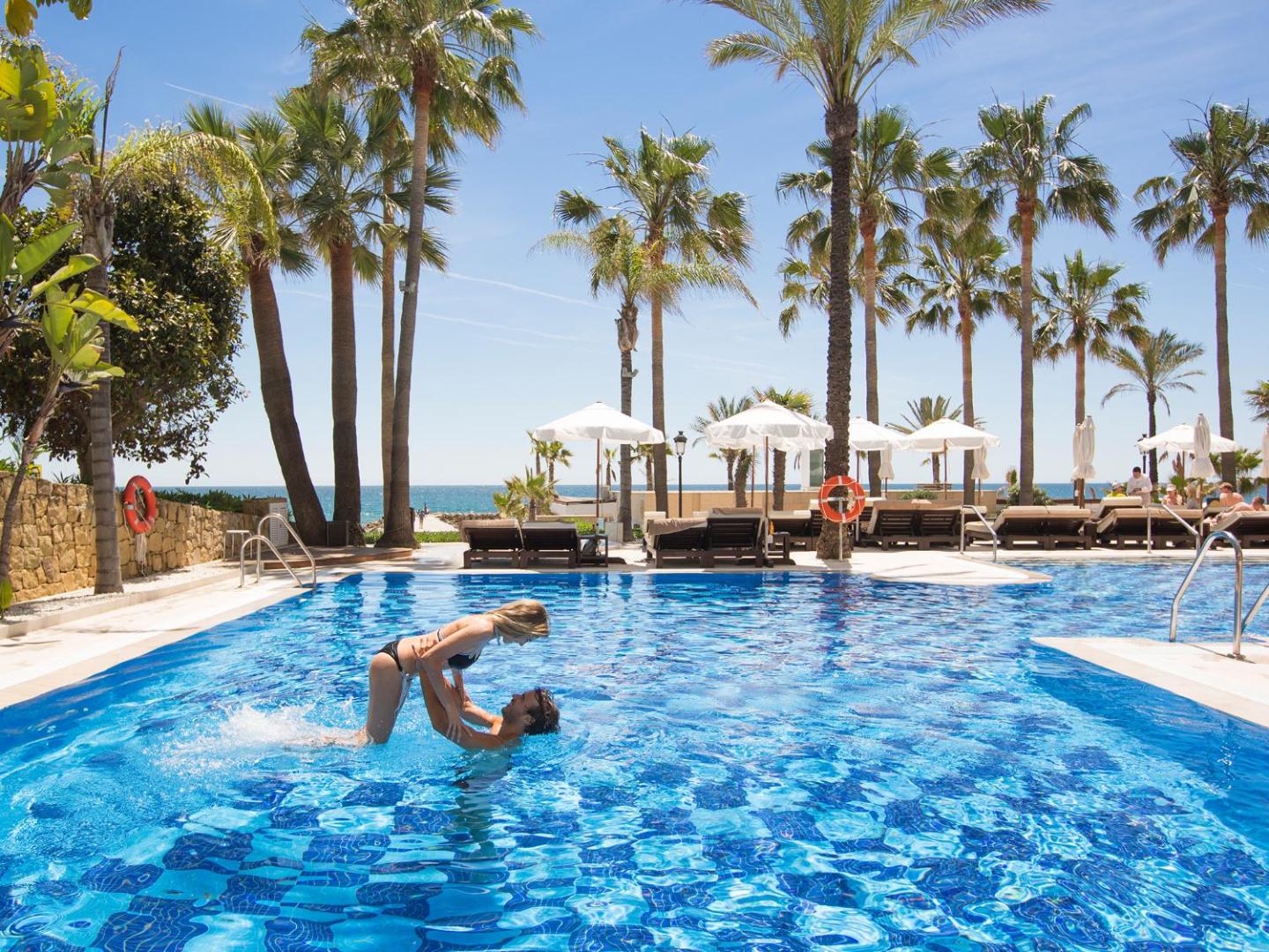 Pāris atpūšas Amare Beach Hotel Marbella āra baseinā, fonā redzamas palmas un balti saulessargi piekrastē