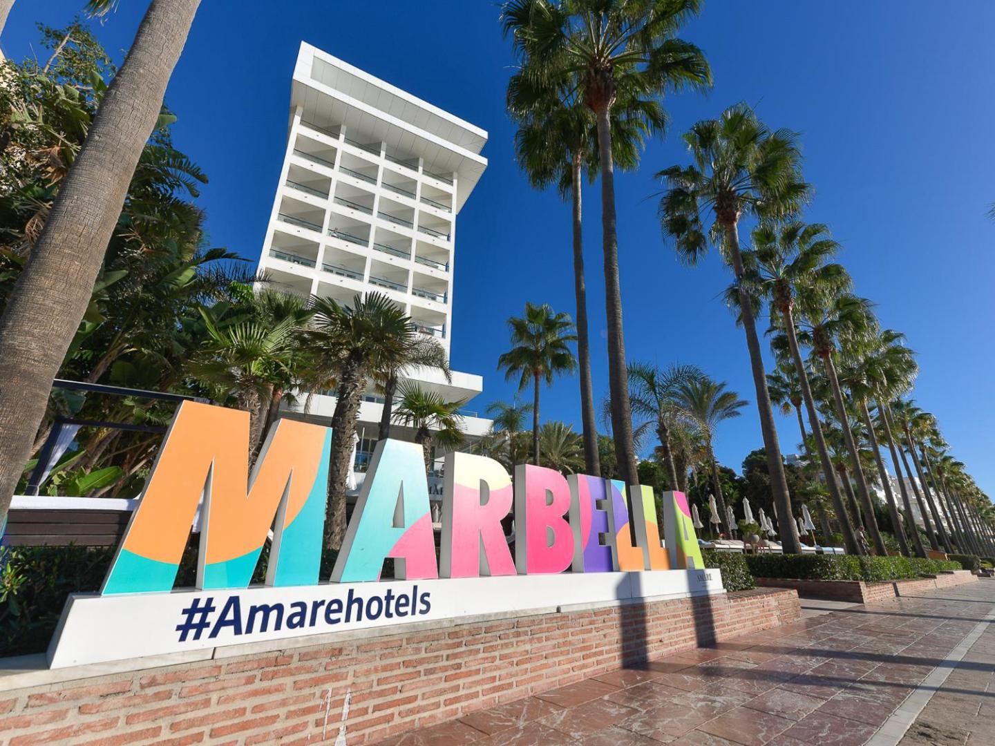 Krāsains Marbella uzraksts pie Amare Beach Hotel Marbella ieejas ar augstām palmām un zilu debesu fonu