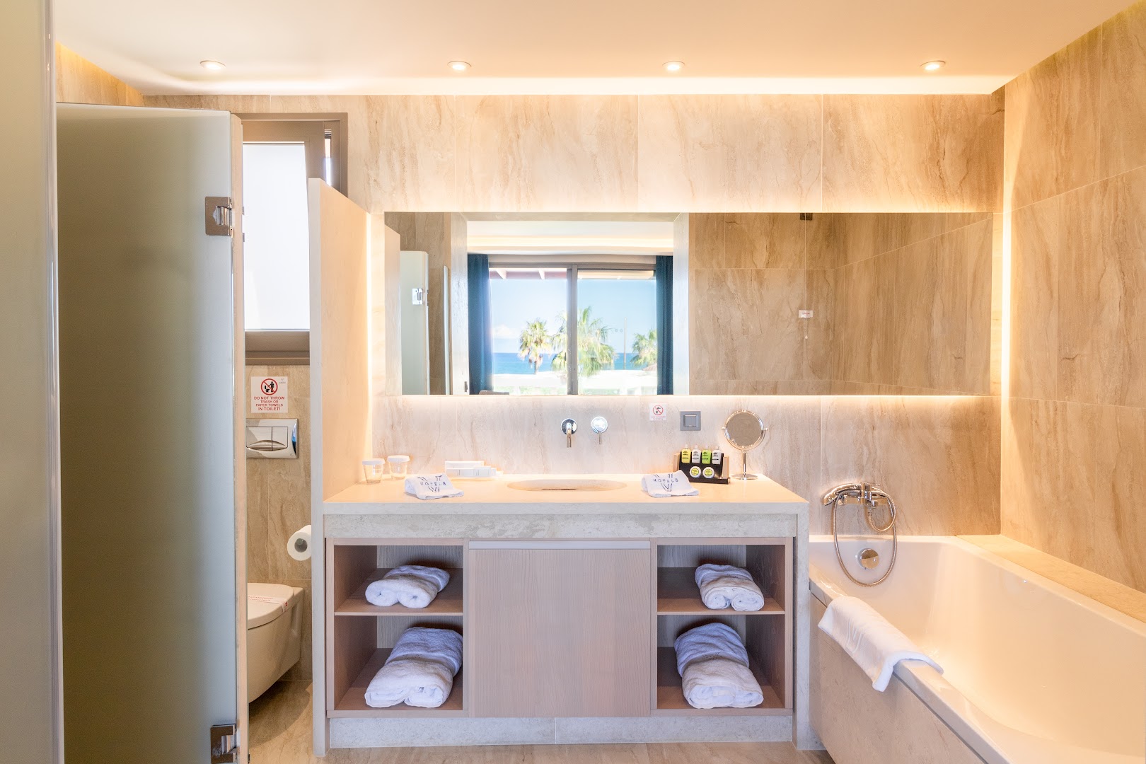 Almyros Beach Resort & Spa numura vannas istaba ar vannu, lielu spoguli, gaišām flīzēm un skatu uz palmām caur logu