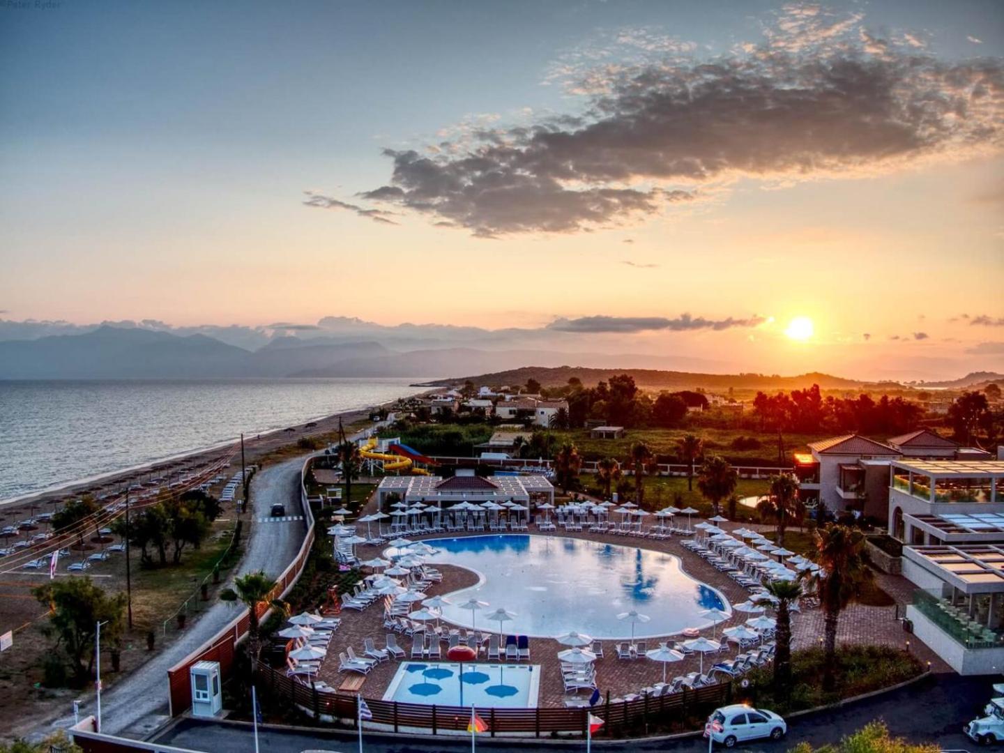 Almyros Beach Resort & Spa panorāmas skats saulrietā Korfu salā — izgaismots baseins, pludmales līnija un kalni tālumā vakara gaismā
