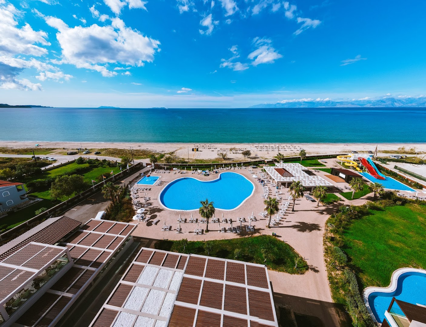 Almyros Beach Resort & Spa centrālais baseins ar saulessargiem un skatu uz Jonijas jūru Korfu ziemeļu piekrastē saulainā dienā