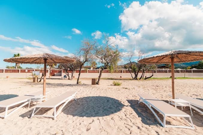 Almyros Beach Resort & Spa pludmales atpūtas zona ar salmu saulessargiem un moderniem sauļošanās krēsliem uz gaišām smiltīm