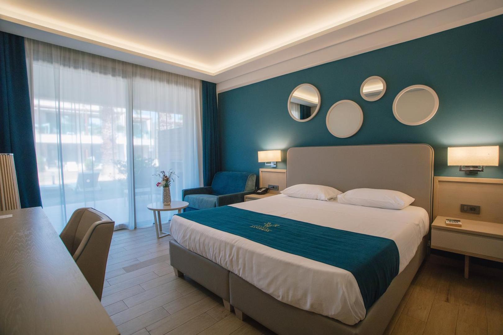 Almyros Beach Resort & Spa moderns divvietīgs numurs ar tirkīza sienu, apaļiem spoguļiem un lielu logu uz terasi Korfu salā