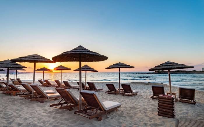 Saulriets High Beach Resort pludmalē Krētā — koka sauļošanās krēsli un salmu saulessargi vakara gaismā pie mierīgas jūras