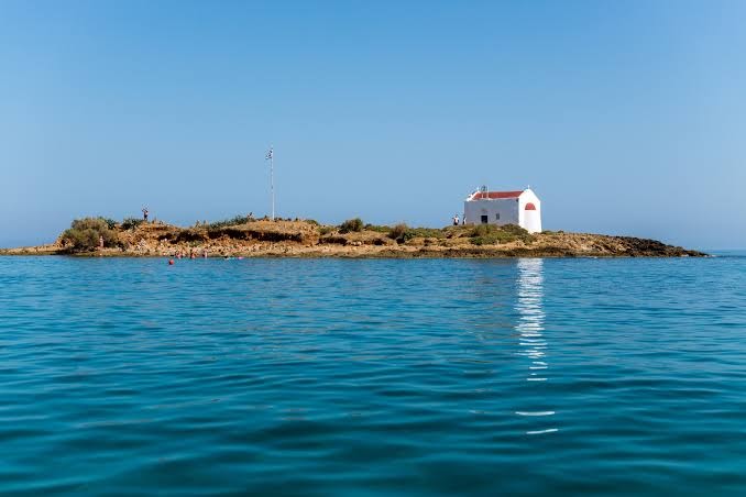 Skats no jūras uz mazu salu ar baltu grieķu baznīcu netālu no High Beach Resort kūrorta Malijā, Krētā