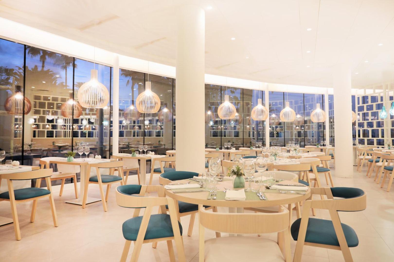 Iberostar Sabila moderns restorāns ar gaišām koka mēbelēm, dizaina lampām un lieliem panorāmas logiem vakarā