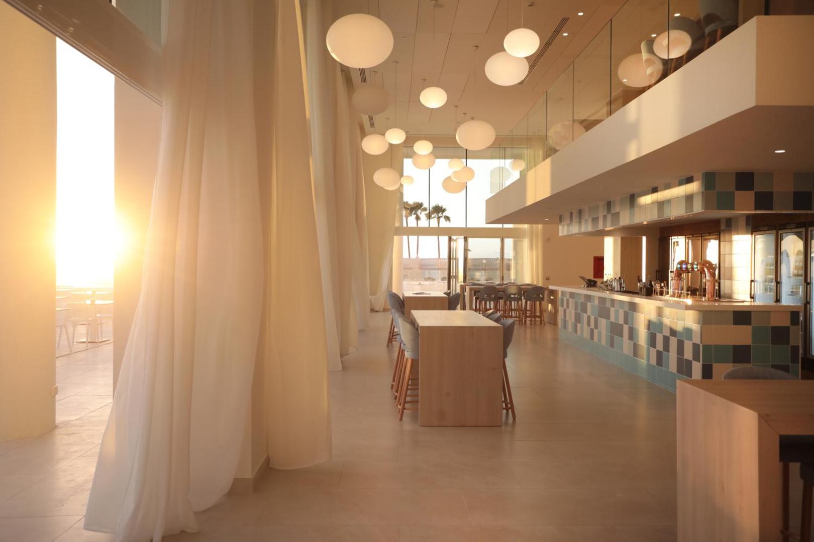 Iberostar Sabila plašais vestibila bārs ar augstiem griestiem un panorāmas logiem saulrieta gaismā, skats uz palmām Tenerifē.