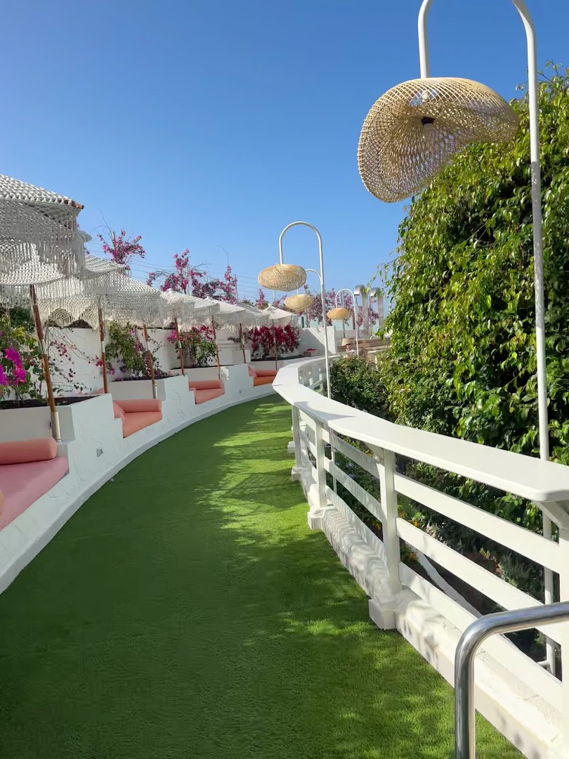 Iberostar Sabila viesnīcas āra terase Tenerifē ar mākslīgo zālienu, rozā atpūtas dīvāniem, dekoratīviem saulessargiem un pītiem gaismas ķermeņiem saulainā dienā.