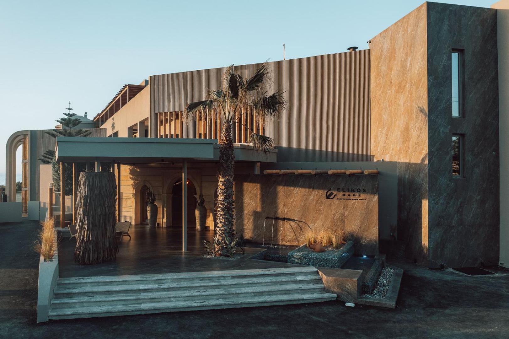 Eliros Mare Beachfront Poem Hotel fasāde dienas gaismā ar dekoratīvu ūdens sienu un modernu arhitektūru Krētas salā