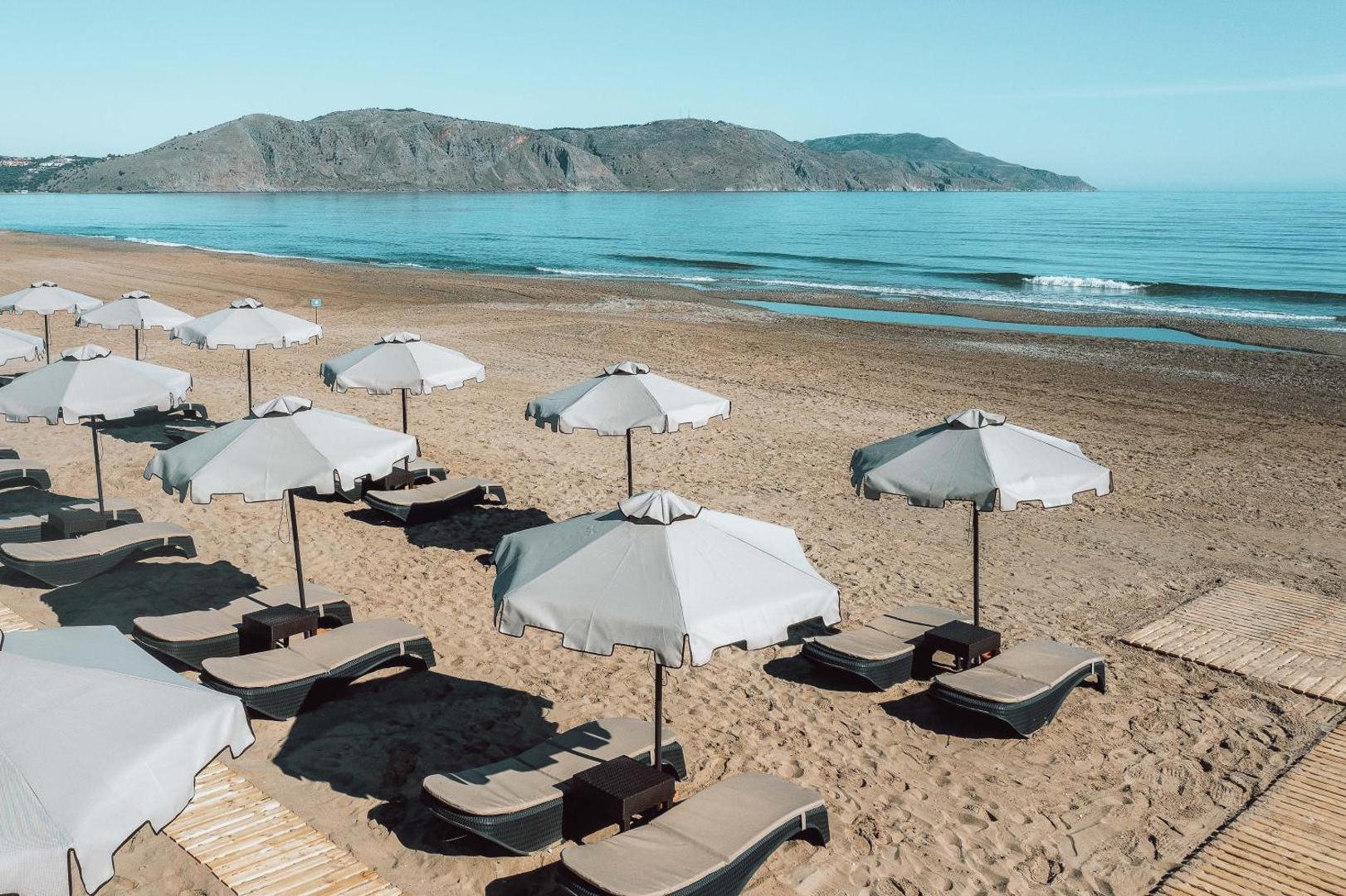 Eliros Mare Beachfront Poem Hotel pludmales skats no sāna — saulessargi un zviļņi uz smilšu fona ar kalniem tālumā