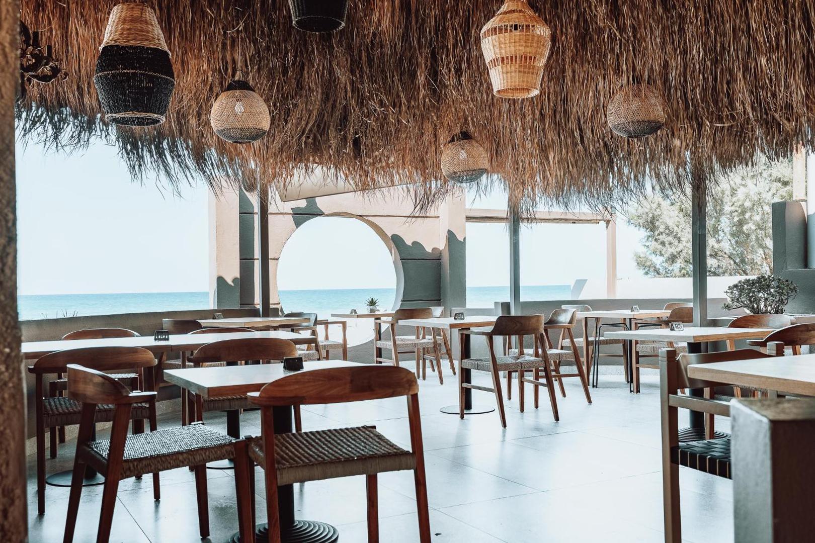 Eliros Mare Beachfront Poem Hotel restorāna terase ar dizaina gaismekļiem, koka mēbelēm un skatu uz jūru caur arkas logu