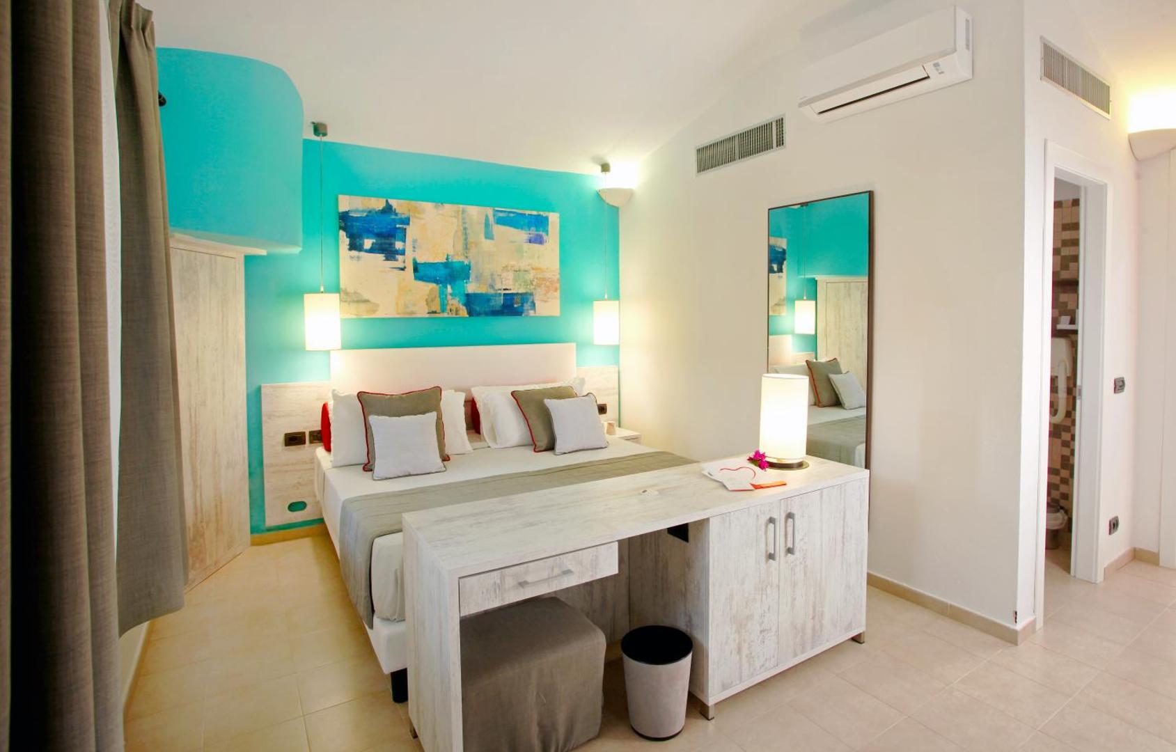 VOI Floriana Resort gaišs divvietīgs numurs ar tirkīza sienu, modernu mākslas darbu, rakstāmgaldu un baltām mēbelēm Kalabrijas piekrastē.