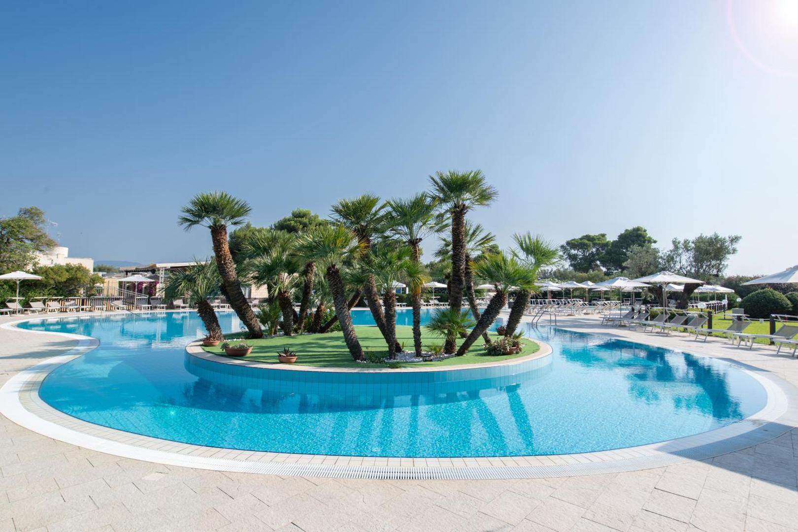 VOI Floriana Resort āra baseins ar palmu salu centrā Kalabrijā, Itālijā — apkārt izvietoti sauļošanās krēsli un saulessargi