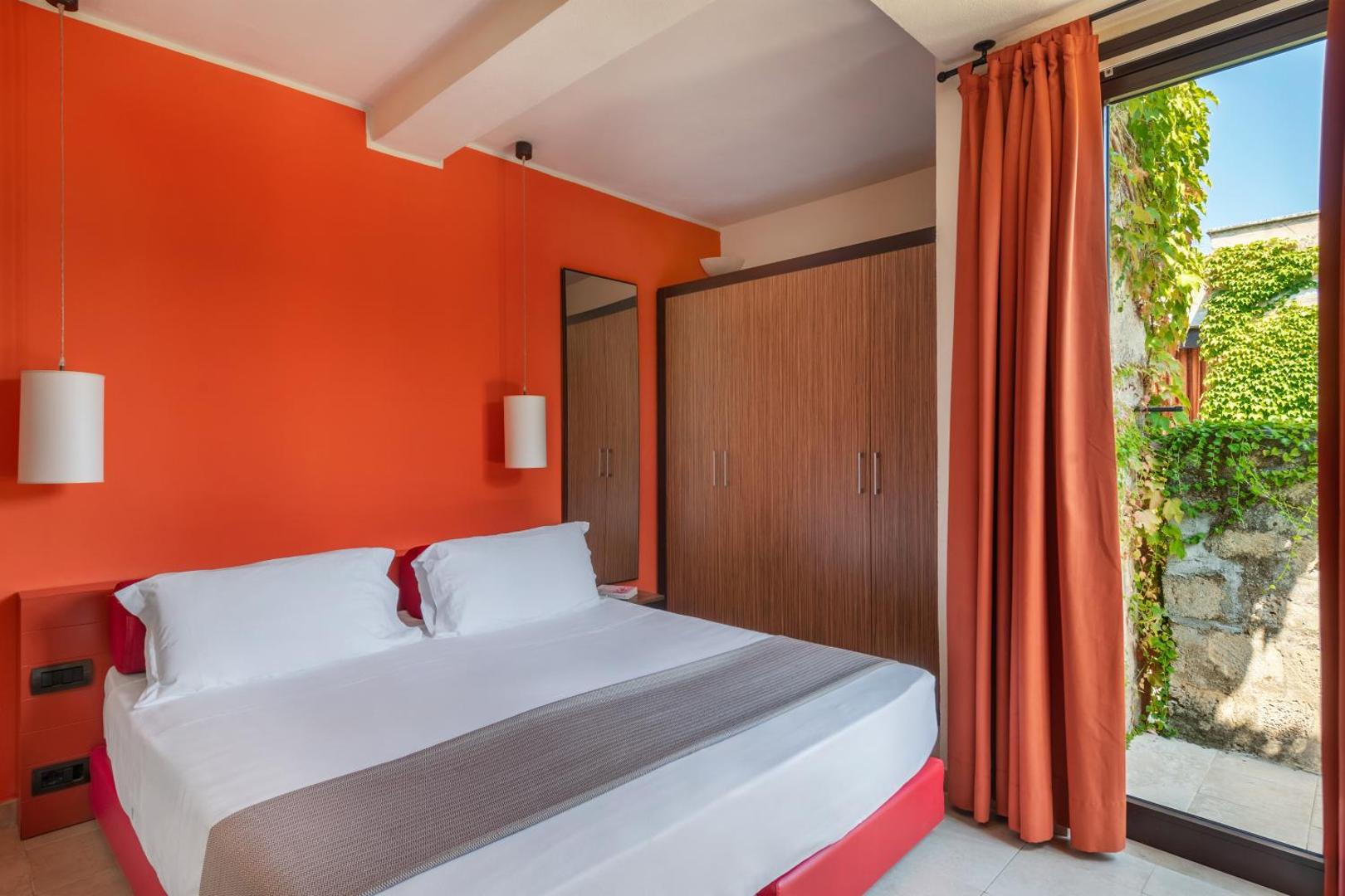 VOI Floriana Resort numurs ar koši oranžu sienu, divguļamo gultu un izeju uz nelielu, apzaļumotu terasi Itālijas kūrortā.